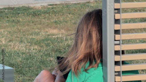 Unrecognizable female student in green shirt sitting on wooden bench outdoors using smartphone to browse social media with hair blowing in wind close up rear view shot on sunny summer day in park