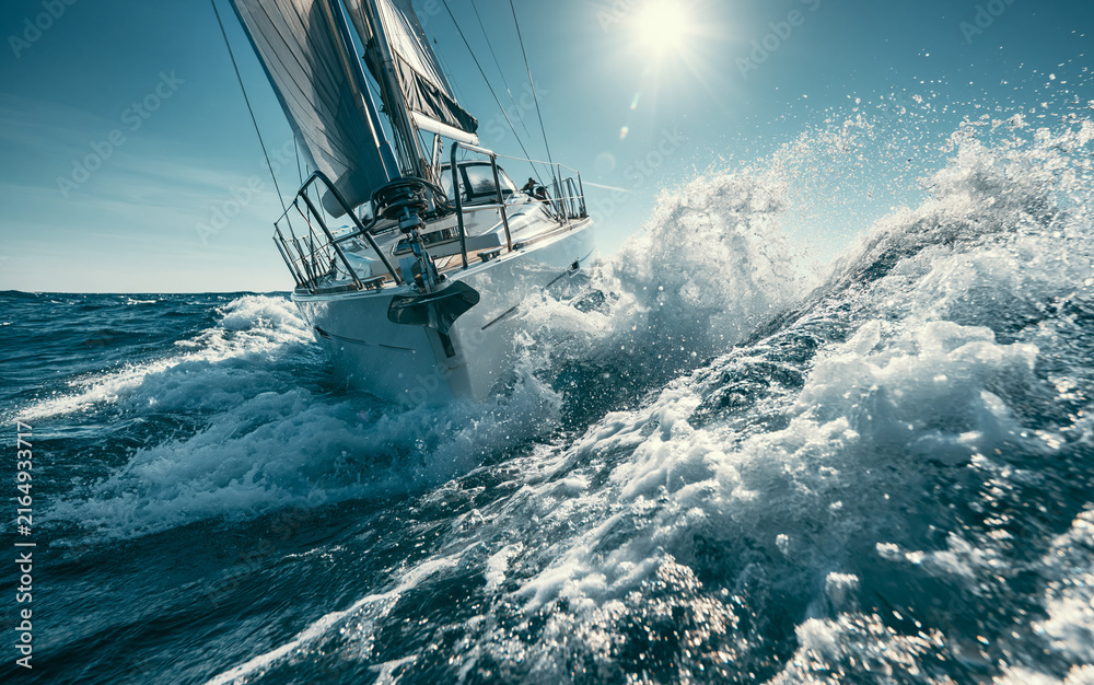 Obraz premium Sleek sailboat races through rough ocean waves. Water splashes high, creating white foam. Bright sun shines on blue sea. Dynamic yacht sailing fast during summer day.
