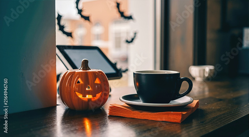 Ceramic jack o lantern pumpkin with a lit tealight next to a black espresso cup. Moody Halloween decoration on a wooden cafe counter