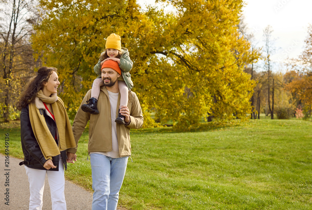 Fototapeta premium Family in autumn park, parents with child daughter. Father carries the girl on shoulders as they walk a path amid golden leaves, sharing happiness and bright smile. Warm family joy and unity.