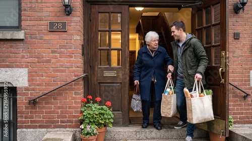 Young man helps elderly woman carry groceries up steps to her apartment building