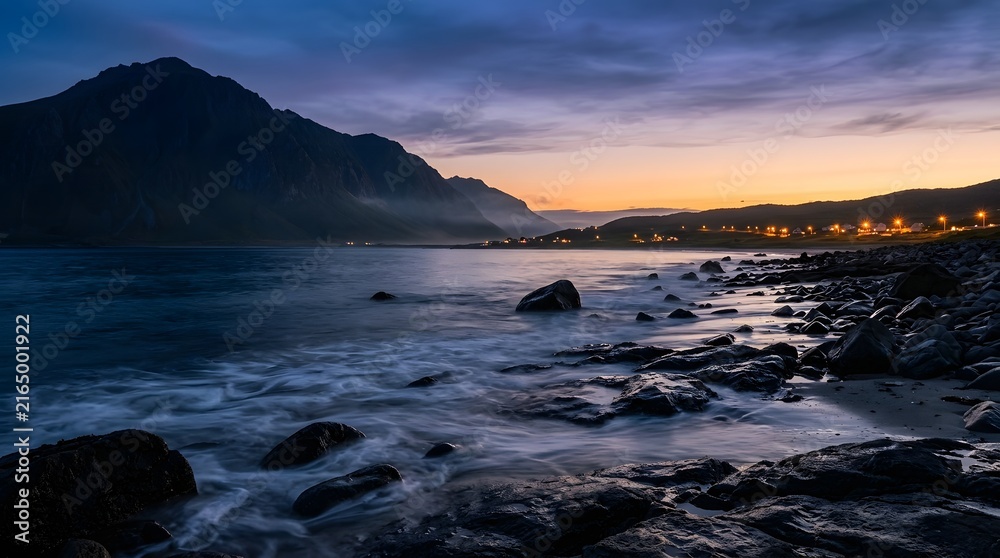 Obraz premium Long exposure landscape of a rocky coast at dusk with mountains, ocean waves, and distant village lights under twilight sky