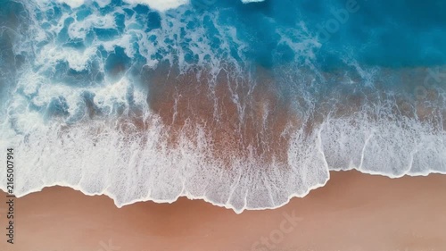 An aerial top-down view shows blue ocean waves crashing onto a sandy beach with white foam.