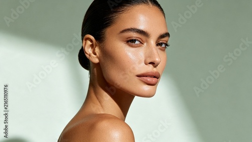 Close-up portrait of a woman with radiant skin and thick eyebrows, looking over her shoulder against a soft green background.