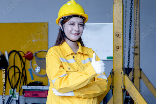 Принти на полотні Portrait of beautiful engineer woman with helmet in yellow safety clothing standing and giving thump up at manufacturing factory
