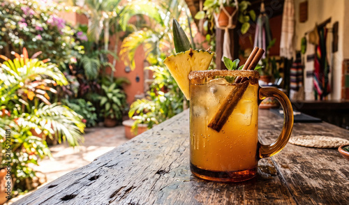 A rich amber glass mug of traditional Tepache on a rustic wooden bar counter, Fermented pineapple drink with ice cubes, garnished with a pineapple wedge, cinnamon stick, and brown sugar rim