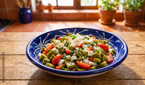 Ensalada de Nopales in a blue hand-painted Talavera bowl on a wooden tabletop, Tender diced tender cactus pads (nopales) tossed with plum tomatoes, white onions, cilantro, queso fresco, and extra virg