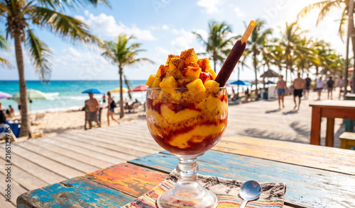 A rich layered Mangonada sorbet dessert in a clear dessert glass on a colorful wooden table, Bright yellow mango sorbet swirled with red chamoy sauce, topped with diced fresh mango chunks, Taj�n chili