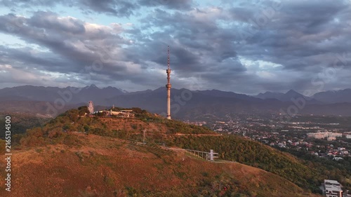 The famous TV tower on Kok Tobe Mountain in the Kazakh city of Almaty, set against a mountain range on a cloudy summer evening.