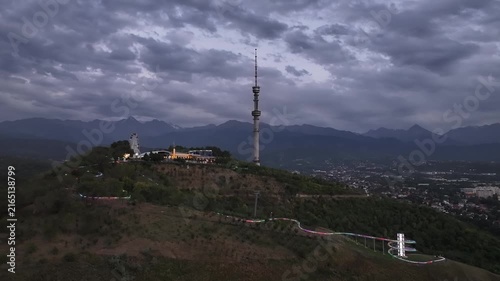 The famous TV tower on Kok Tobe Mountain in the Kazakh city of Almaty, set against a mountain range on a cloudy summer evening.