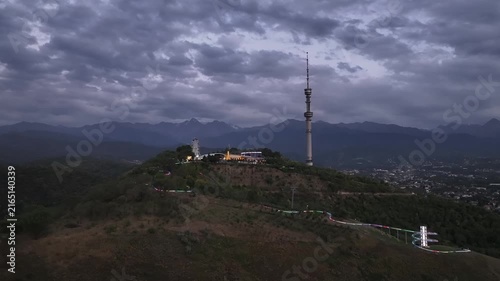 The famous TV tower on Kok Tobe Mountain in the Kazakh city of Almaty, set against a mountain range on a cloudy summer evening.