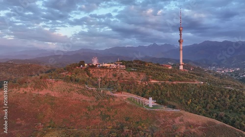 The famous TV tower on Kok Tobe Mountain in the Kazakh city of Almaty, set against a mountain range on a cloudy summer evening.