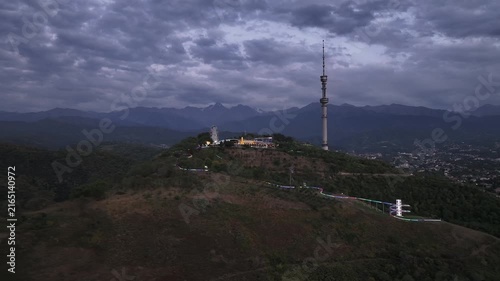 The famous TV tower on Kok Tobe Mountain in the Kazakh city of Almaty, set against a mountain range on a cloudy summer evening.