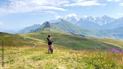 Woman riding electric bike through flowering alpine meadow with mountain peaks
