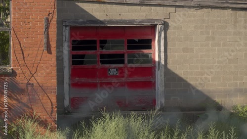 Slow zoom-out shot of an abandoned factory with a red garage door. Broken windows. Destroyed industrial building. Creepy.