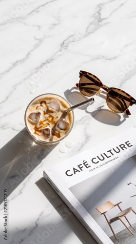 Overhead flat lay of iced caramel macchiato on marble table with sunglasses and harsh sunlight shadows.