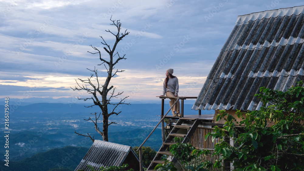 Fototapeta premium A woman sitting on wooden balcony, looking at mountain views on cloudy morning