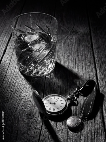 Black and white still life of a vintage pocket watch and a glass of iced liquor on a rustic wooden table