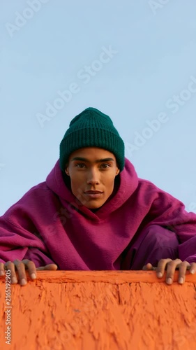 Person in bright magenta hoodie and green beanie leaning over an orange ramp against clear blue sky for portrait background and banner design