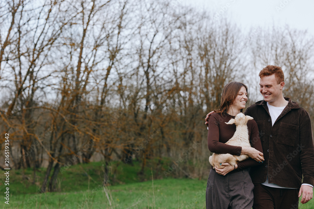 Naklejka premium Happy couple outdoors with a dog, smiling and affectionate, walking through a park in bright daylight, enjoying nature and each other, expressing warmth and companionship on a calm day.