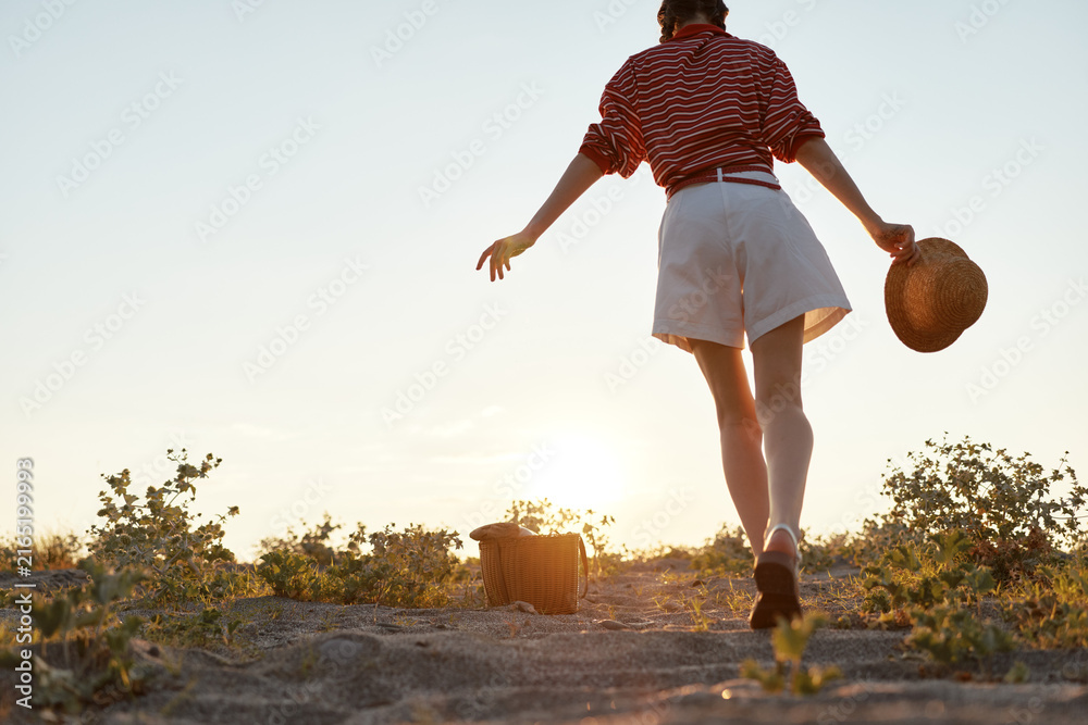 Fototapeta premium A young woman in a striped shirt and white shorts walks toward a golden sunset on a sandy path, her hat swinging in a relaxed, carefree summer moment.
