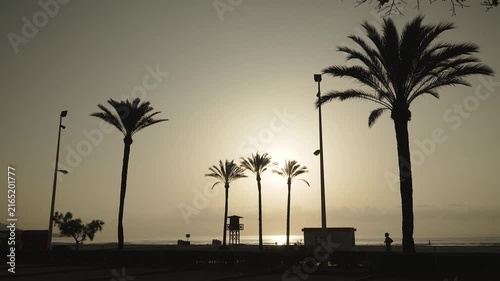 Silhouettes of palms, beach infrastructure against hot scorching rising sun. Silhouette of runner jogging on sandy beach during morning, wide shot. Sports activity in cool time during extreme heat.