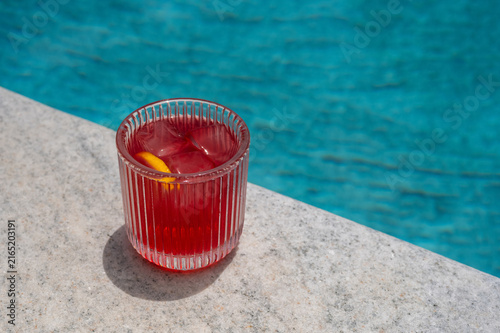 Red cocktail with ice and lemon slice by the swimming pool edge.
