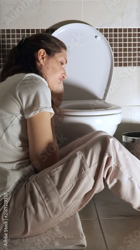 Sick young woman feeling unwell with nausea and stomach ache sitting on the bathroom floor, experiencing morning sickness symptoms of pregnancy, food poisoning, or alcohol intoxication