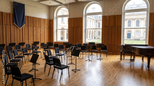 Music academy rehearsal hall with chairs, music stands, grand piano, wooden floor, and large arched windows