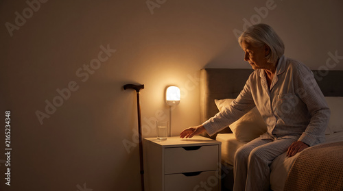 Elderly woman in pajamas reaching for bedside light in dim bedroom with walking cane nearby, calm atmosphere