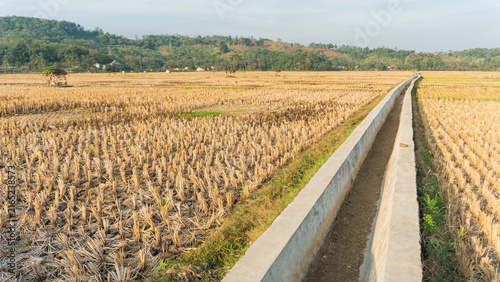 A rural farmland after harvest and a dried-up irrigation canal during the dry season, set against a backdrop of the sky, a small shelter, and mountains.