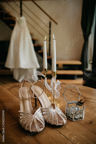 Accessories for the bride on her wedding day in a hotel room: wedding dress nearby, festive shoes, box with gold wedding rings, candles in candelabras on the table