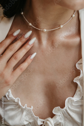 Bride in a white dress with a necklace around her neck holds her hand near her face showing off the white manicure on her fingers on her wedding day