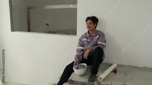 Construction inspector in gray uniform sitting on concrete floor holding white safety helmet smiling near construction materials at unfinished building site showing worker safety and rest moments