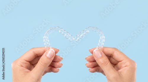 Close-up of female hands holding two clear plastic orthodontic aligners forming a heart shape against a pastel blue background. Perfect for modern aesthetic dentistry and invisible braces advertising.