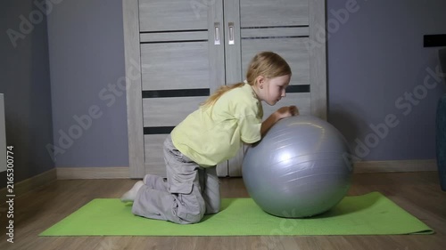Little caucasian girl on green mat exercising at home with fitness ball during day. Child performing physical activity on wooden floor of living room. Healthy lifestyle development.