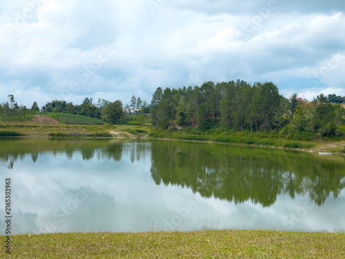 Serene lake with lush green trees and cloudy sky reflecting on the water surface.