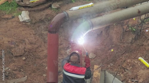 Skilled worker welding large metal pipes in an excavated trench, bright sparks flying from the joint, construction process of heating main infrastructure, industrial scene slow motion
