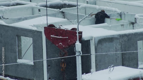 Person clearing snow from modular concrete structures, red crane hook in foreground, heavy snowfall creating winter scene at construction site during cold day