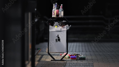 Outdoor garbage can overflowing with empty plastic glass soda beer bottles, night outside store entrance, waste disposal problem. Pollution concept