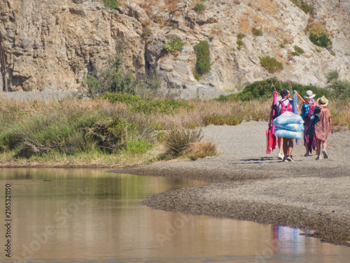 Bild auf Leinwand Group of tourists carrying beach gear walking along sandy shore of Laghetti di Marinello natural reserve lagoon with limestone rock wall backdrop in Oliveri, Tindari, Sicily, Italy