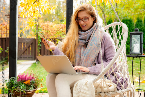A mid adult woman works remotely on a laptop while drinking tea in a hanging swing chair on an autumn patio.