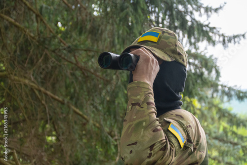 Quadro su tela Masked soldier scanning forest with binoculars and Ukrainian flag patches