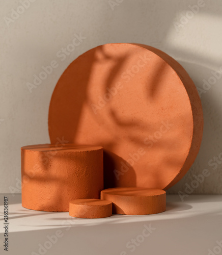 Three terracotta textured cylinder product display podiums  arranged in front of a large terracotta round disc on a beige wall background with leaf shadow overlay.