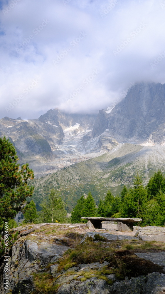 Fototapeta premium Banc en pierre face à un paysage alpin composé de conifères, de sommets rocheux et d’un glacier partiellement couvert par les nuages à Chamonix-Mont-Blanc, dans les Alpes françaises.