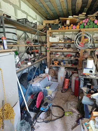 Highly cluttered workshop interior with tools and machinery. Disorganized garage storage with shelving units and painted logs