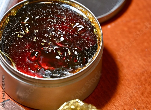 A commercial close-up photograph of an open round metal tin filled with dark, solid rosin flux used for electronics soldering and electrical work. The hard, amber-colored resin surface glistens under