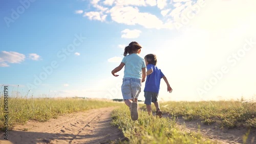 Two children, a girl and a boy, run together along a dirt path. The girl and the boy enjoy a sunny day in a grassy field.