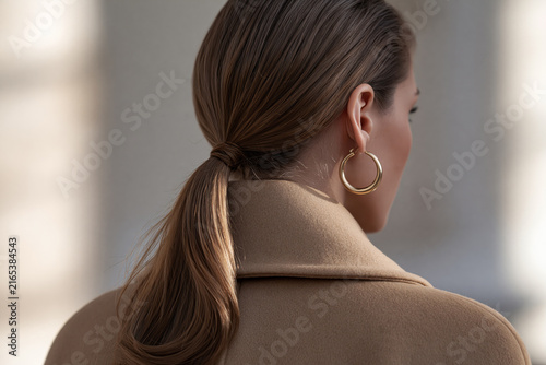 Rear view of woman with sleek low ponytail, hair tie, gold hoop earring and camel wool coat collar.