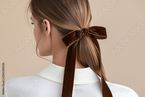 Woman with ponytail tied with brown velvet ribbon bow, white blouse on beige background.